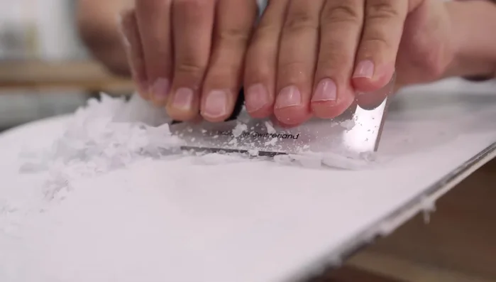 Scrape off excess wax with a plastic scraper held at a 45° angle, working from nose to tail in one direction. Use the notch on the scraper for edges. Tackle stubborn areas with the more rigid end of the scraper.